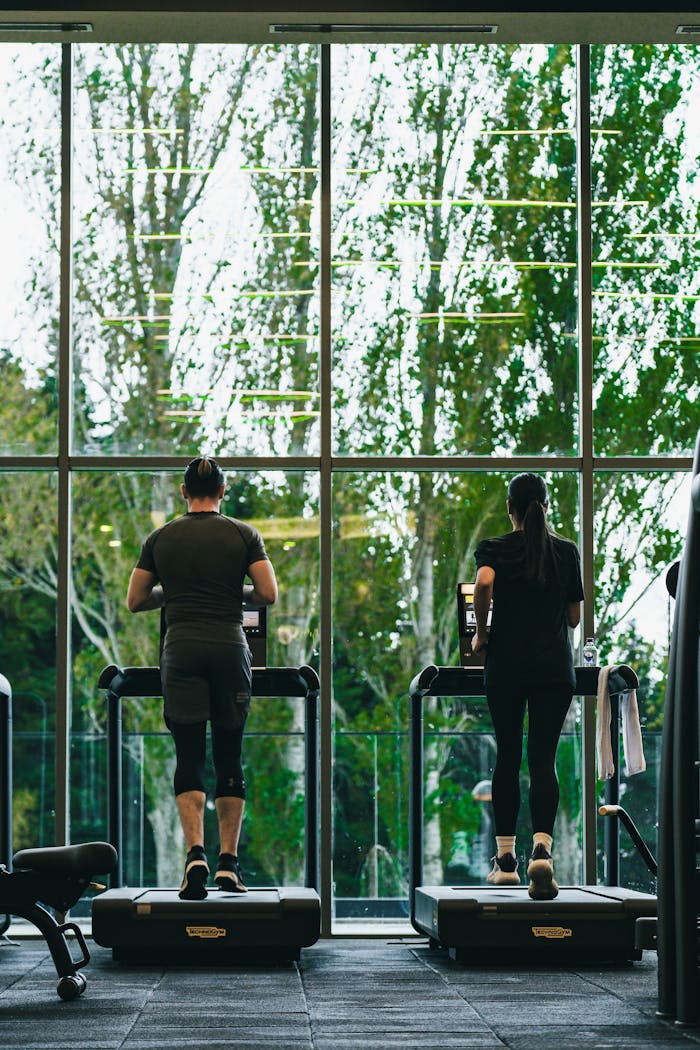 Two fitness enthusiasts running on treadmills in a gym with outdoor view.