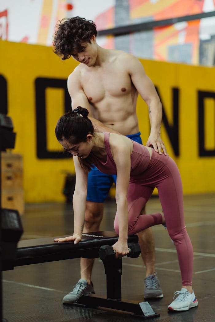 A personal trainer assists a woman in a gym workout with weights, focusing on proper form.
